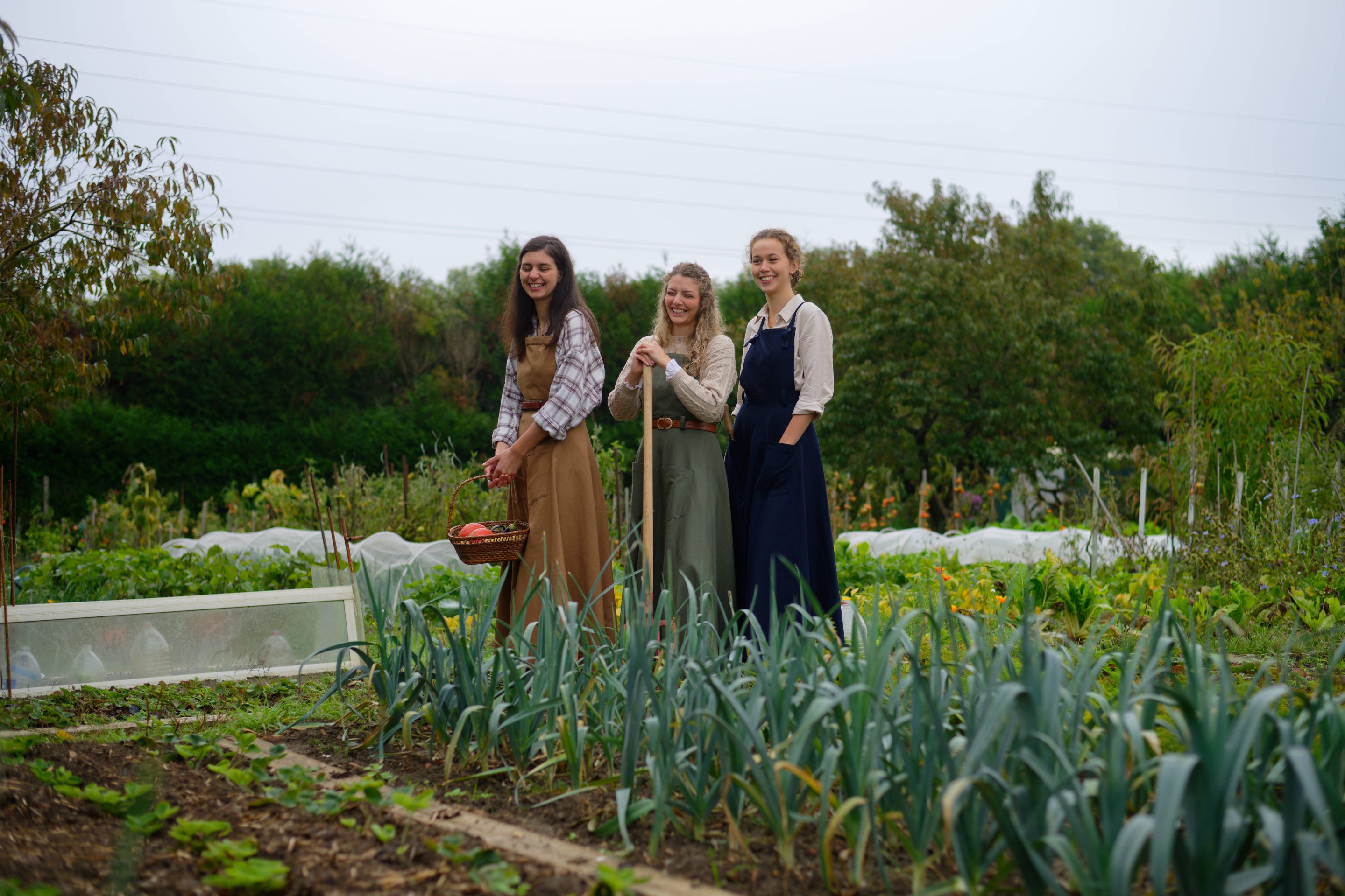 Trois femmes souriantes en vêtements de travail féminin, la robe-salopette, dans un potager